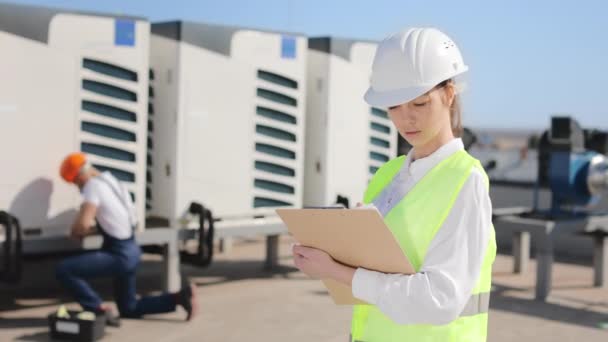 Portrait of a happy woman engineer is making entries in documents. The engineer is repairing the air conditioning system in the background. They are on the roof of a business center. They are wearing — Vídeo de Stock