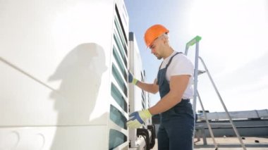 The engineer is making an inspection of the air conditioning system. He is checking the external state. He is wearing work gloves and work clothes. He is on the roof of a business center. Nice sunny