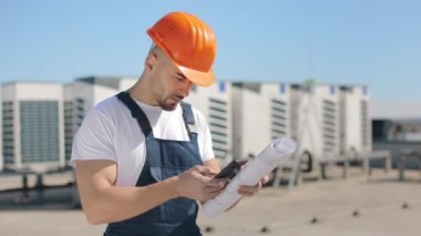 A serious engineer is writing data to a smartphone and looking at the air conditioning system. He is holding a drawing of the project. He is wearing work clothes and a hard hat. Nice sunny day. 4K