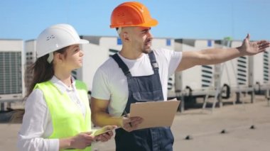 Two engineers are discussing the work done to install the air conditioning system. The engineer is pointing his hand at the object. They are wearing work clothes and a hard hat. Air conditioning