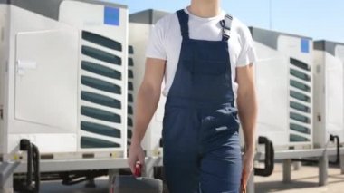A happy engineer is going and holding a toolbox in his hands. He is wearing work clothes and a hard hat. Hes on the roof of a business center. Air conditioning systems in the background. Nice sunny