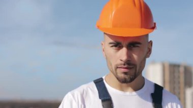 Portrait of a serious engineer who is looking at the camera. He is on the roof of a business center. He is wearing work clothes and a hard hat. Construction in the background. Nice sunny day. 4K