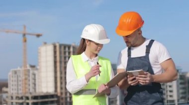 Two young engineers are discussing working documents. A man is holding a tablet. They are wearing work clothes and a hard hat. They are on the roof of a business center. Construction in the background