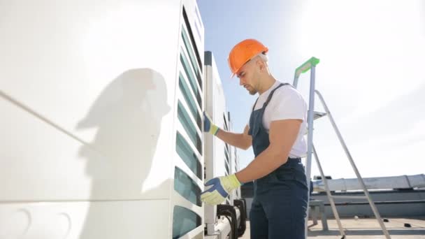 The engineer is making an inspection of the air conditioning system. He is checking the external state. He is wearing work gloves and work clothes. He is on the roof of a business center. Nice sunny — Wideo stockowe