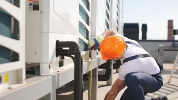 The engineer is using a tape measure to measure parts of the air conditioner and checking the distances between them. He is wearing work clothes and a hard hat. Nice sunny day. 4K — Wideo stockowe