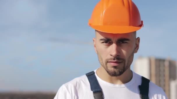 Portrait of a serious engineer who is looking at the camera. He is on the roof of a business center. He is wearing work clothes and a hard hat. Construction in the background. Nice sunny day. 4K — Wideo stockowe