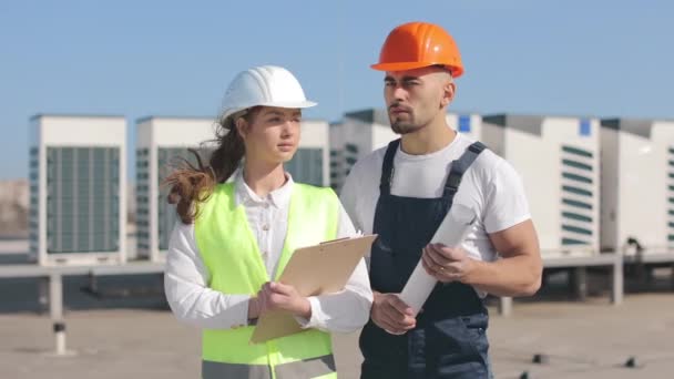 Two happy engineers are standing and watching the camera. They are on the roof of a business center. They are wearing work clothes and a hard hat. Air conditioning systems in the background. Nice — Stock Video