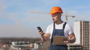 Genç mühendis akıllı telefonuyla mesaj atıyor. Odaklanmış ve elinde bir dosya dolusu belge var. O, iş kıyafetleri ve sert bir şapka giyiyor. Bir iş merkezinin çatısında. İnşaat...