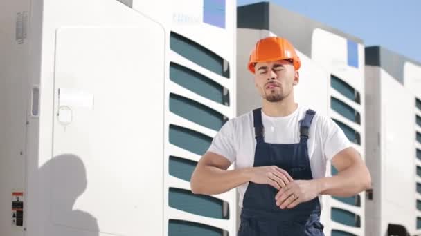 Portrait d'un jeune ingénieur qui croise les bras et regarde la caméra. Il porte des vêtements de travail et un casque. Systèmes de climatisation en arrière-plan. Il est sur le toit d'une entreprise — Video