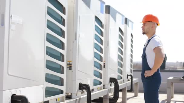 A seriously young engineer is standing and looking at the air conditioning system. He is keeping his hands on his belt. He is wearing work clothes and a hard hat. Hes on the roof of a business center — Stockvideo