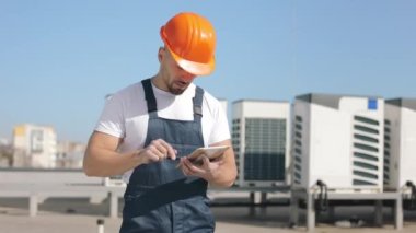 Ciddi bir genç mühendisin portresi tablet üzerinde çalışıyor ve kameraya bakıyor. Bir iş merkezinin çatısında. O, iş kıyafetleri ve sert bir şapka giyiyor. Klima sistemi çalışıyor.