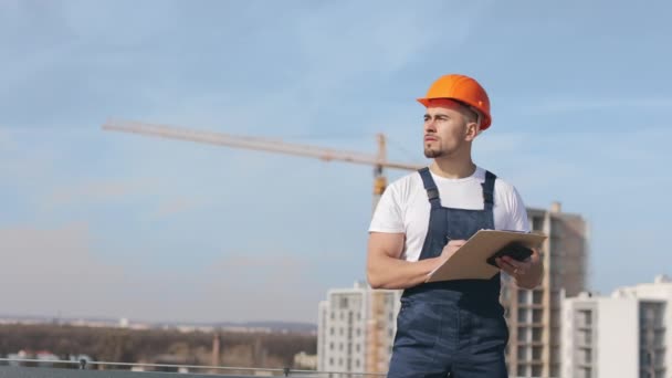 Porträt eines jungen Ingenieurs, der auf dem Dach eines Geschäftszentrums steht. Er schaut in die Unterlagen und macht sich Notizen. Er trägt Arbeitskleidung und einen harten Hut. Bauarbeiten in der — Stockvideo