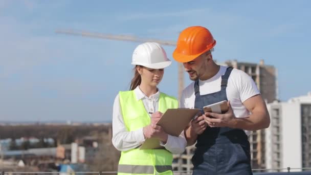 Two young engineers are discussing working documents. They are raising their heads and happy looking at the camera. A man is holding a tablet. They are wearing work clothes and a hard hat. They are on — Stock Video