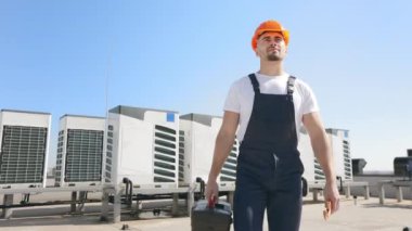 Mutlu bir mühendis elinde bir alet çantası tutuyor. Boru anahtarını ellerinin arasında tutuyor. O, iş kıyafetleri ve sert bir şapka giyiyor. Bir iş merkezinin çatısında. Hava