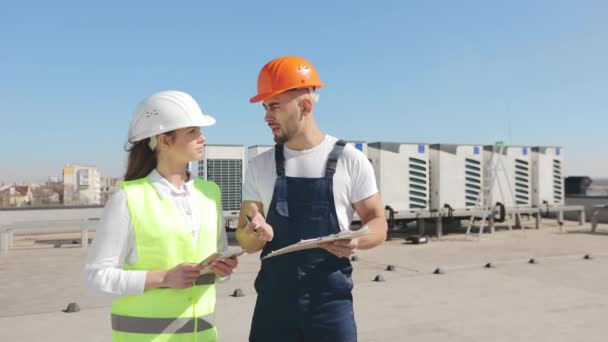 Two engineers are discussing an air conditioning system project. They are holding documents and a tablet in their hands. They are on the roof of a business center. They are wearing work clothes and a — Wideo stockowe