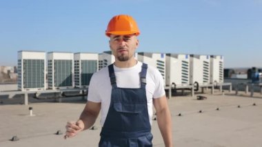 Mühendis duygusal olarak konuşuyor ve kameraya bakıyor. Havalandırma sistemini işaret ediyor. Bir iş merkezinin çatısında. O, iş kıyafetleri giyiyor. 4K