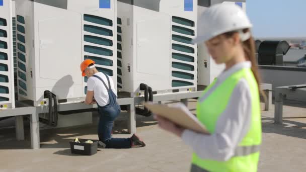 The young concentrated engineer is maintaining the air conditioning system. He is turning the screw with a pipe wrench. A female engineer is checking his work and standing in the foreground. They are — Stock Video