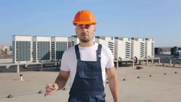 L'ingegnere sta parlando emotivamente e guardando la telecamera. Sta indicando l'impianto di condizionamento. E 'sul tetto di un centro commerciale. Indossa abiti da lavoro. 4K — Video Stock