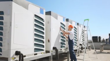 Mühendis havalandırma sistemini teftiş ediyor. O, dış durumu kontrol ediyor. O, iş kıyafetleri ve sert bir şapka giyiyor. Bir iş merkezinin çatısında. Güzel güneşli