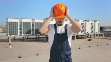 Kafasına koruyucu kask takan genç bir mühendisin portresi. O ciddi ve kameraya bakıyor. O, iş kıyafetleri giyiyor. Arka planda havalandırma sistemi var. 4K