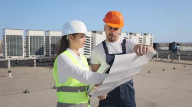İki mühendis proje çizimlerini tartışıyor. Ellerinde belgeler ve çizimler var. Bir iş merkezinin çatısındalar. İş kıyafetleri ve kalın bir şapka giyiyorlar. Hava