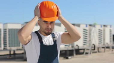 Kafasına koruyucu kask takıp ellerini kemerine koyan mutlu bir mühendisin portresi. O, iş kıyafetleri giyiyor. Arka planda havalandırma sistemi var. Çatıda.