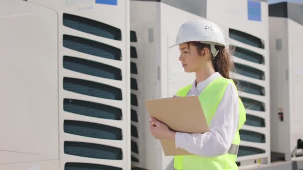 Portrait of a young woman who is standing and making notes. She is looking after the engineers work. She is on the roof of a business center. She is wearing work clothes and a hard hat. Nice sunny — Stock Video