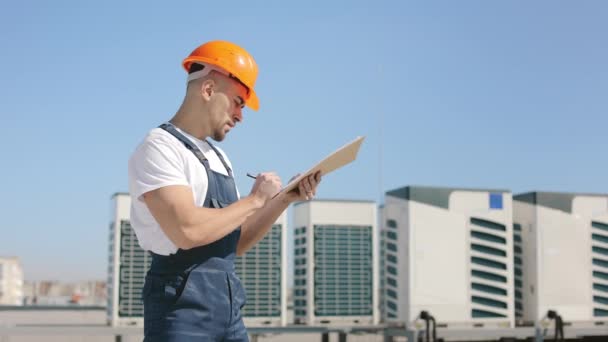 Młody inżynier bada system klimatyzacji i wprowadza dane do dokumentów. Ma na sobie ubranie robocze i twardy kapelusz. Jest na dachu centrum biznesowego. Ładny słoneczny dzień. — Wideo stockowe