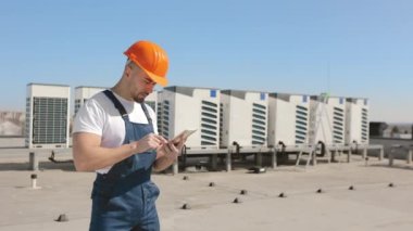 Ciddi bir genç mühendisin portresi tablet üzerinde çalışıyor ve havalandırma sistemlerine bakıyor. Bir iş merkezinin çatısında. O, iş kıyafetleri ve sert bir şapka giyiyor. Güzel güneşli