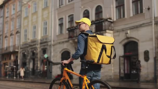 Ένας χαρούμενος ντελιβεράς περπατάει στο κέντρο της παλιάς πόλης και κρατάει ένα ποδήλατο μαζί του. Κουβαλάει ένα κίτρινο σακίδιο για παράδοση στους ώμους του. Πυροβολισμός από πίσω. 4K — Αρχείο Βίντεο