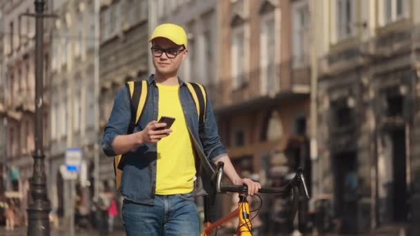Młody, szczęśliwy dostawca idzie i pisze smsy na smartfonie. Trzyma ze sobą rower. Ma przy sobie żółty plecak, który ma dostarczyć na ramionach. Centrum starego miasta. 4K — Wideo stockowe