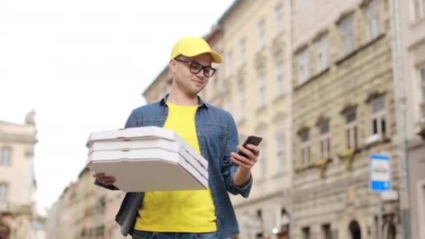 Ένας νεαρός χαρούμενος ντελιβεράς στέκεται και κρατάει τρία κουτιά πίτσας. Χαμογελάει και στέλνει μηνύματα στο smartphone. Φοράει κίτρινο καπέλο και γυαλιά. Το κέντρο της παλιάς πόλης στο βάθος. 4K — Αρχείο Βίντεο