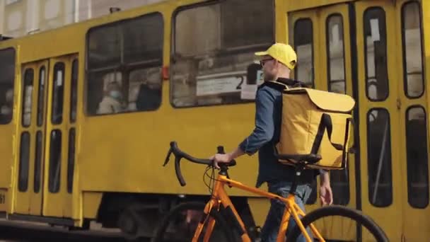 Um homem de parto feliz está andando pelo centro da cidade velha e segurando uma bicicleta com ele. Ele está carregando uma mochila amarela para entrega em seus ombros. O eléctrico está a passar em segundo plano. 4K — Vídeo de Stock