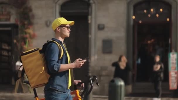 Een jonge vrolijke bezorger gaat en houdt een smartphone vast. Hij heeft een fiets bij zich. Hij draagt een gele rugzak voor levering op zijn schouders. De camera is vanaf de zijkant aan het opnemen. 4K — Stockvideo