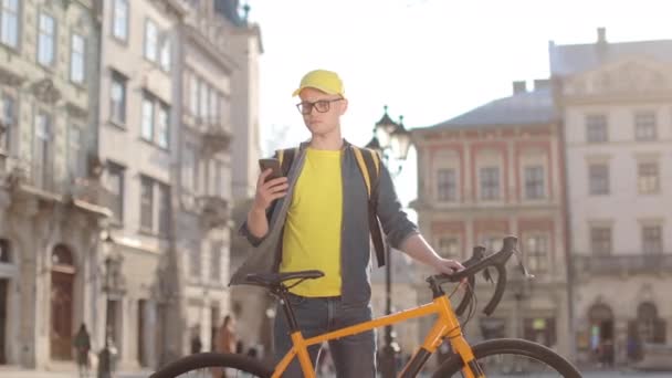 Portrait d'un livreur heureux qui se tient debout et envoie des SMS sur un smartphone. Il tient un vélo. Il porte un sac à dos jaune sur ses épaules. La caméra se déplace de gauche à droite. 4K — Video