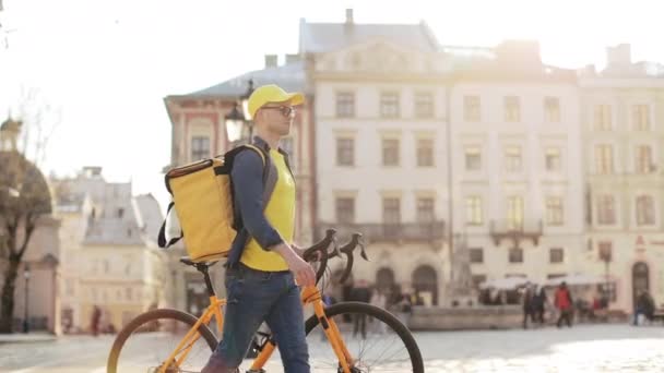 Un repartidor feliz que va y sostiene una bicicleta. Lleva ropa amarilla. Lleva una mochila amarilla para entregarla en sus hombros. Disparando desde el costado. 4K — Vídeo de stock