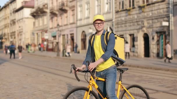 Portret van een vrolijke bezorger die op een fiets zit en naar de camera kijkt. Hij draagt gele kleding. Hij draagt een gele rugzak op zijn schouders. Centrum van de oude stad. 4K — Stockvideo