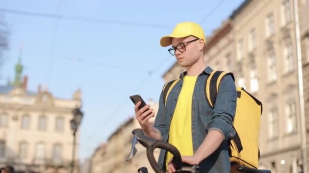Retrato de um homem de entrega feliz que está sentado em uma bicicleta e mensagens de texto no smartphone. Ele está carregando uma mochila amarela para entrega em seus ombros. A câmera está se movendo da esquerda para a direita. 4K — Vídeo de Stock