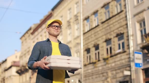 Молодий щасливий чоловік з доставкою їде і тримає три коробки для піци. Він посміхається. Він у жовтій шапці та окулярах. Центр старого міста на задньому плані. 4-кілометровий — стокове відео
