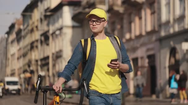Πορτρέτο ενός νεαρού ντελιβερά που πηγαίνει και κρατά ένα smartphone. Κρατάει ένα ποδήλατο μαζί του. Κουβαλάει ένα κίτρινο σακίδιο για παράδοση στους ώμους του. Στο κέντρο της παλιάς πόλης. 4K — Αρχείο Βίντεο