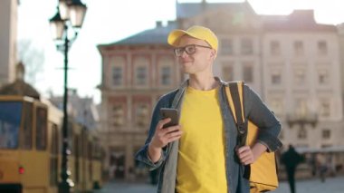 Akıllı telefondan mesaj atan mutlu bir teslimatçının portresi. Sarı bir şapka ve gözlük takıyor. O, omuzlarında sarı bir sırt çantası taşıyor. Tramvay geliyor. 4K