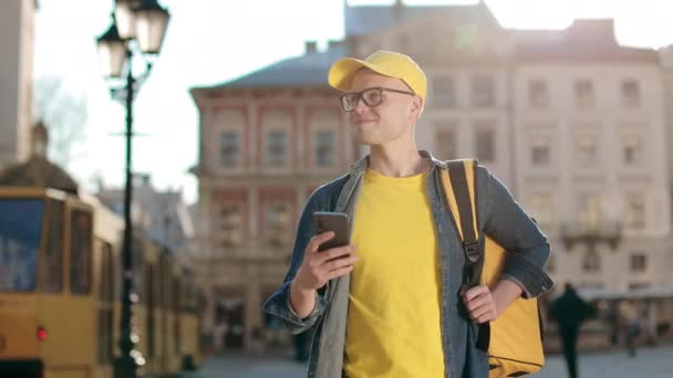 Portrait d'un jeune livreur heureux qui va envoyer des SMS sur le smartphone. Il porte une casquette jaune et des lunettes. Il porte un sac à dos jaune sur ses épaules. Un tram roule. 4K — Video