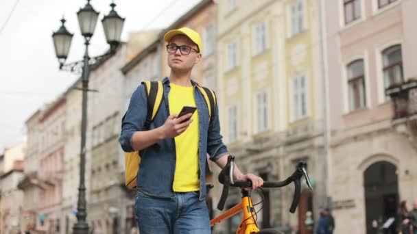 Mladý poslíček jde a píše smartphone. Drží s sebou kolo. Má na sobě žluté šaty. Nosí na ramenou žlutý batoh k porodu. 4K — Stock video