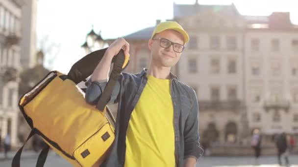 Portret młodego, szczęśliwego kuriera, który stoi i wyrzuca plecak przez ramię. Ma na sobie żółtą czapkę i okulary. Kamera porusza się od lewej do prawej. 4K — Wideo stockowe