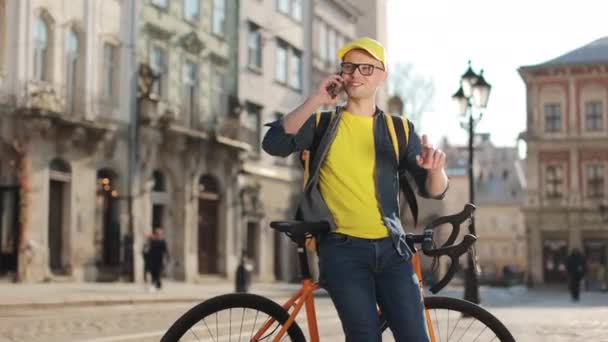 Portrét mladého doručovatele, který sedí na kole a emocionálně mluví po telefonu. Na ramenou má žlutý batoh. Kamera se pohybuje zleva doprava. 4K — Stock video