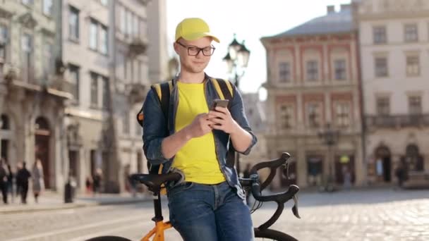 Портрет щасливого чоловіка, який сидить на велосипеді і пише на смартфоні. Він несе жовтий рюкзак для доставки на плечі. Камера рухається зліва направо. 4-кілометровий — стокове відео