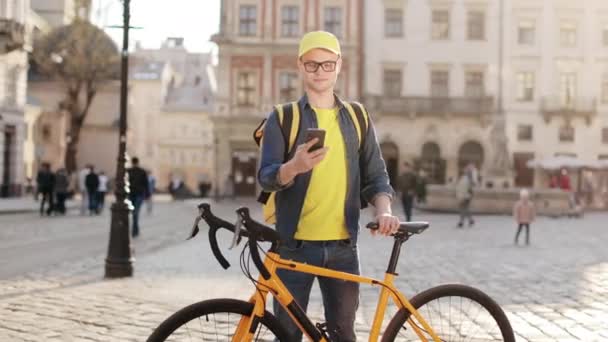 Πορτρέτο ενός διανομέα που στέκεται και στέλνει μηνύματα σε ένα smartphone. Κρατάει ένα ποδήλατο. Φοράει κίτρινο καπέλο και γυαλιά. Κουβαλάει ένα κίτρινο σακίδιο στους ώμους του. 4K — Αρχείο Βίντεο