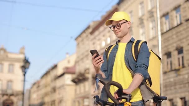 Portrét šťastného doručovatele, který stojí a drží smartphone. Drží v ruce kolo. Na ramenou má žlutý batoh. Kamera se pohybuje zleva doprava. 4K — Stock video