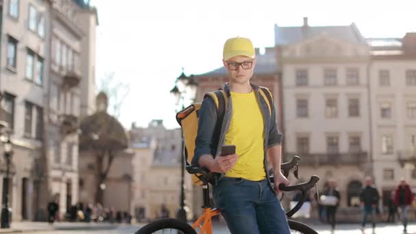 Portret van een jonge bezorger die op een fiets zit en sms 't op een smartphone. Hij draagt een gele rugzak voor levering op zijn schouders. De camera beweegt van links naar rechts. 4K — Stockvideo