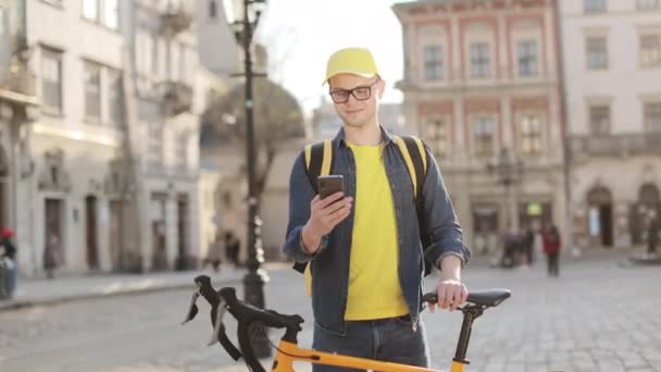 Retrato de um homem de entrega feliz que está de pé e mensagens de texto em um smartphone. Ele está segurando uma bicicleta. Ele tem uma mochila amarela nos ombros. A câmera está se movendo da esquerda para a direita. 4K — Vídeo de Stock